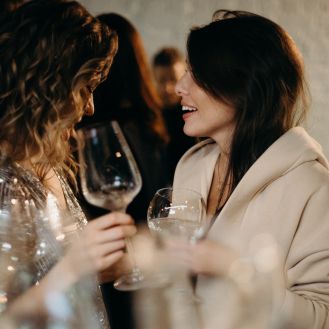 Two elegantly dressed women clink glasses of wine in a festive setting.