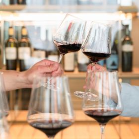 Two hands clinking glasses of wine in front of a fine wine rack.