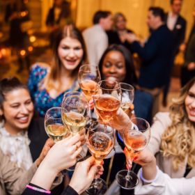 An elegant group of women hold filled wine glasses in the air and clink glasses in festive surroundings.